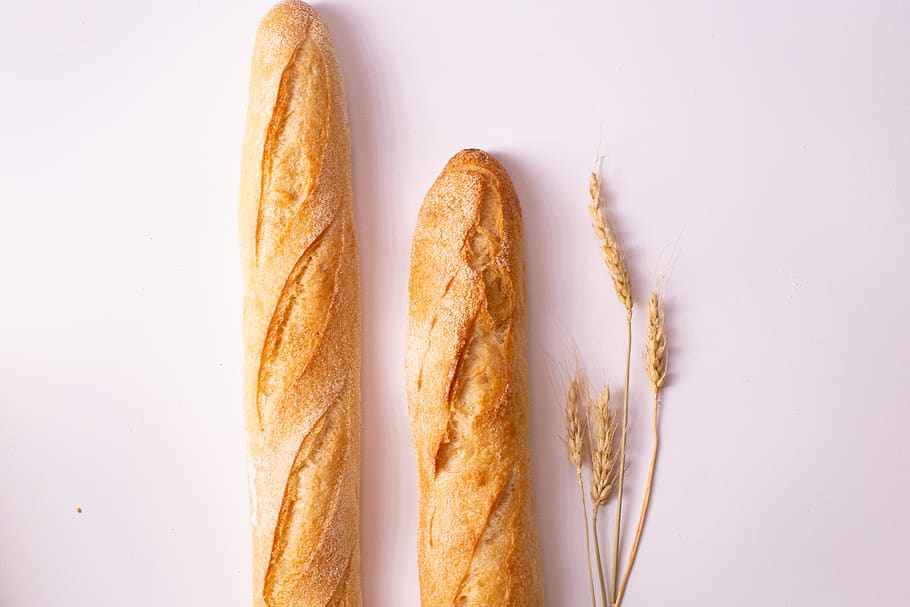 Two French baguettes laying next to wheat on a white table.