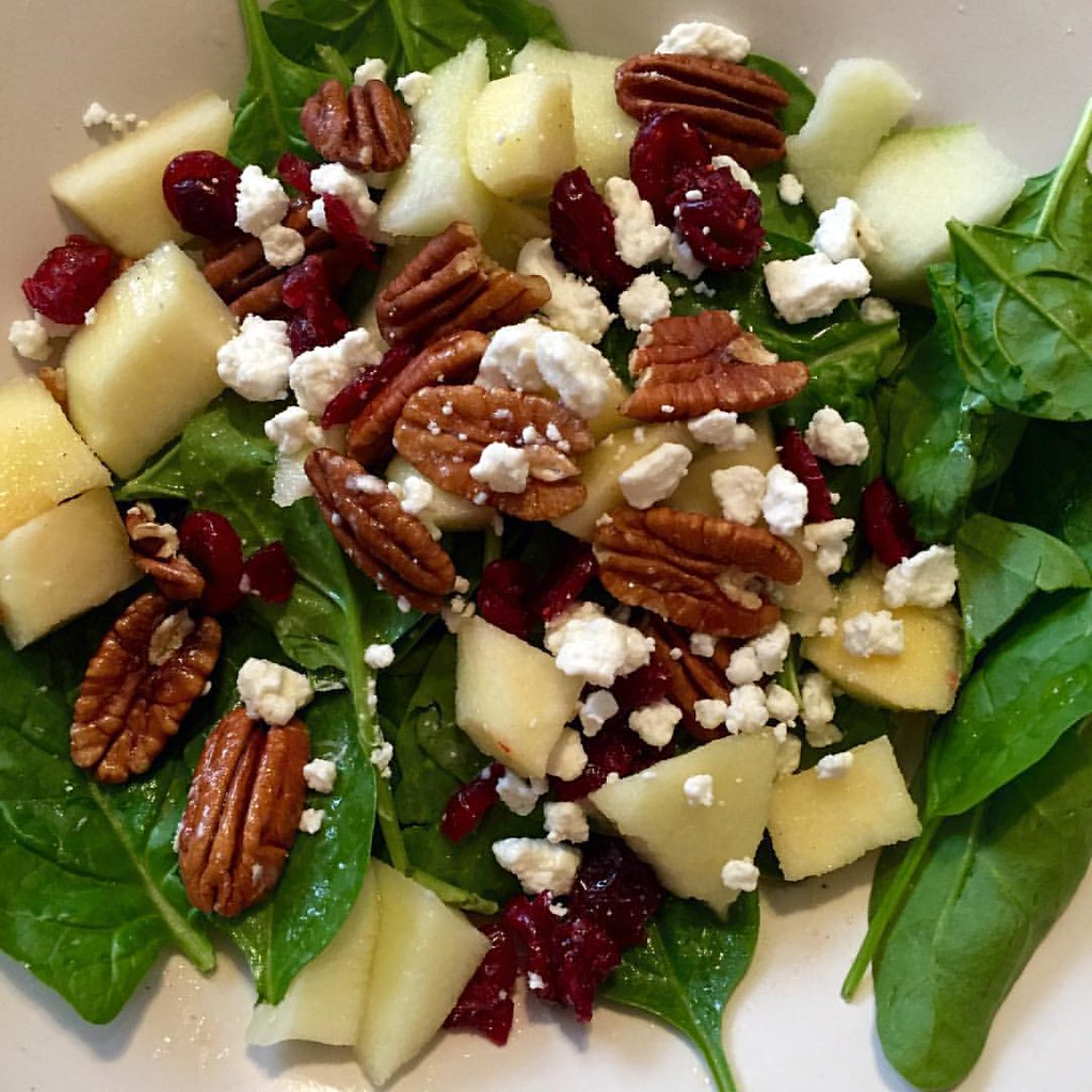 Spinach salad with apples, cranberries, pecans and goat cheese in a white dish.